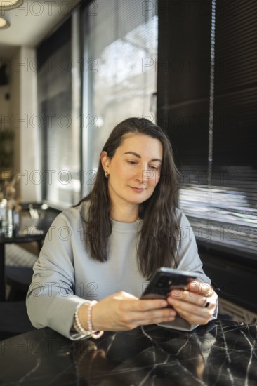 A mid-aged woman focuses intently on her smartphone while sitting at a cafe table bathed in sunlight filtering through the window
