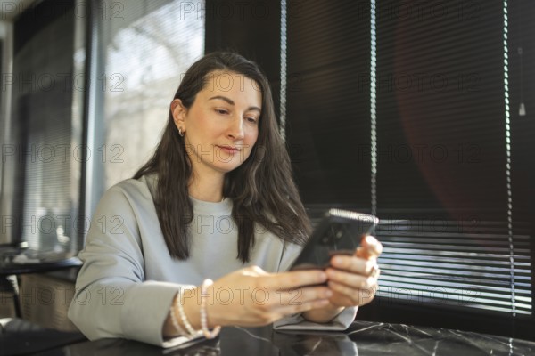 A mid-aged woman is casually using her smartphone in a sunlit cafe environment, reflecting a moment of modern, connected living