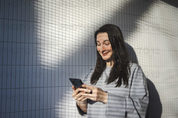 A mid-aged woman enjoys the sunlight as she smiles and engages with her smartphone outdoors against a tiled wall