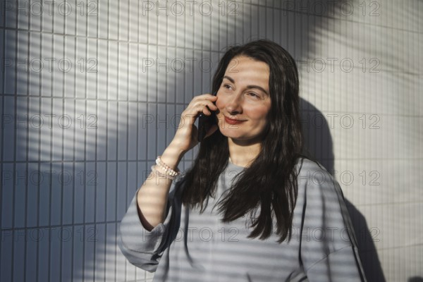 A mid-aged woman enjoys a sunny day at a cafe, chatting on her smartphone against a geometric backdrop