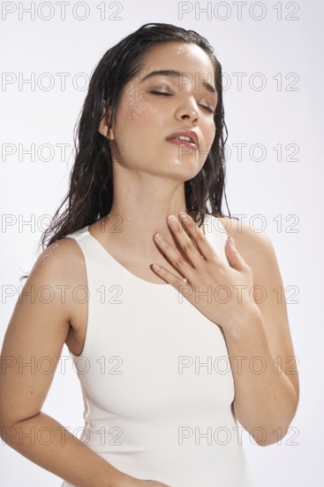 A female model with wet hair and water droplets on her face, wearing a white sleeveless top, poses gracefully, with eyes closed in a studio setting