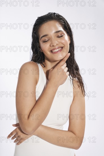 Studio portrait of a joyful female model with wet hair and skin, her eyes gently closed, wearing a white bodysuit, embodying freshness and beauty