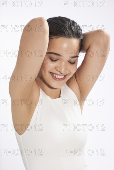 A joyful young female model with wet hair and glitter on her skin, wearing a white dress, looks down while posing in a studio setting