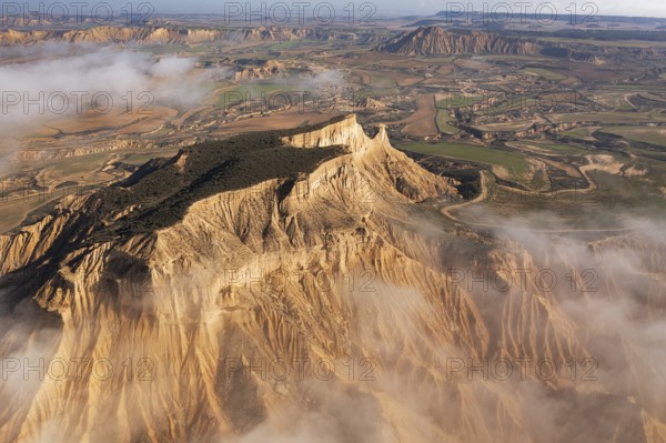 A breathtaking aerial view of rugged mountains partially covered in mist, surrounded by patchwork farmlands stretching into the horizon under a clear sky