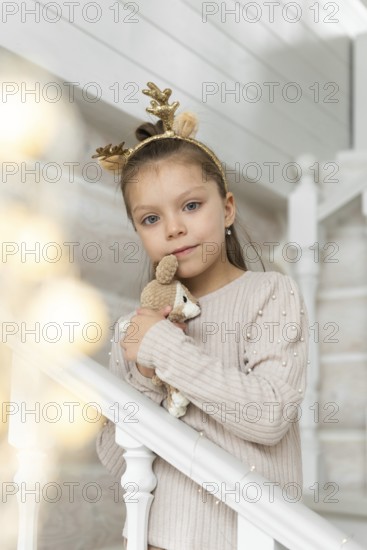 A young girl, dressed in a soft beige sweater adorned with pearls, celebrates Christmas. She wears a golden reindeer antler headband and holds a plush toy. Her blue eyes are looking at the camera, capturing a moment of holiday warmth and innocence