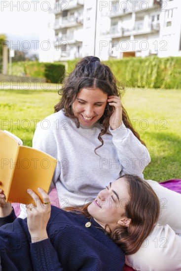 A young women smiles while lying down looking at her partner, who is reading from an orange book in a park. They display warmth and joy against an urban apartment backdrop, capturing a tender lgtbiq+ moment