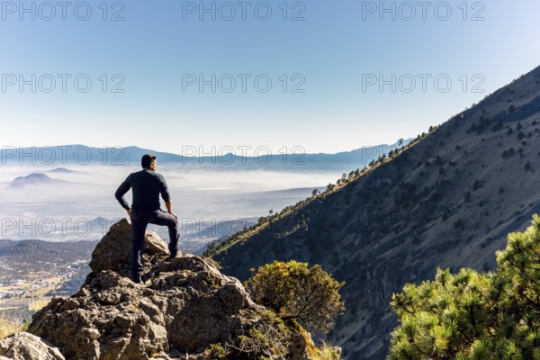 A person stands triumphantly on a rocky mountain peak, gazing over a vast, sunlit valley with distant mountains Clear blue sky and scattered trees create a breathtaking view
