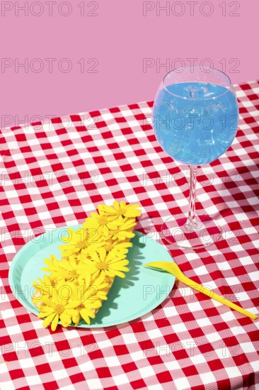 A vibrant summer picnic scene featuring a glass of iced blue beverage and a plate with yellow flowers on a red-checkered tablecloth against a pink backdrop
