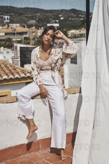 Young woman enjoying a sunny day on a rooftop, dressed in stylish bohemian attire The scenery includes rooftops and distant hills under a bright blue sky