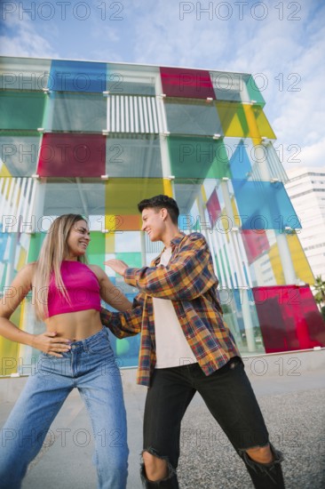 A joyful lesbian couple dances and laughs together in an urban setting. The vibrant glass wall in the background adds a dynamic and artistic touch, symbolizing love, fun, and self-expression