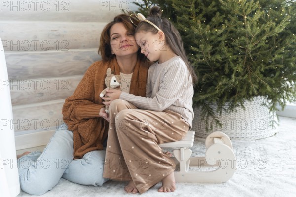 A heartwarming image showing a mother and daughter embracing beside a Christmas tree, capturing the joy and intimacy of family celebrations during the holiday season. Both are smiling gently with eyes closed, enjoying the moment