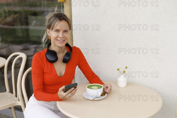 Smiling woman in a bright orange top sits at a Warsaw cafe table She wears headphones around her neck, holding a smartphone, with a matcha latte on the table