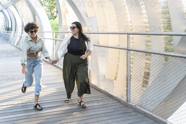 A lesbian couple holds hands as they joyfully walk on a sunlit, modern bridge. Both wearing casual clothes and sunglasses, they share a happy moment in the afternoon light