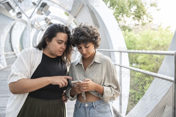 A lesbian couple shares a moment on a contemporary walkway, looking at a smartphone. The scene highlights connection, technology, and urban style amidst a natural backdrop
