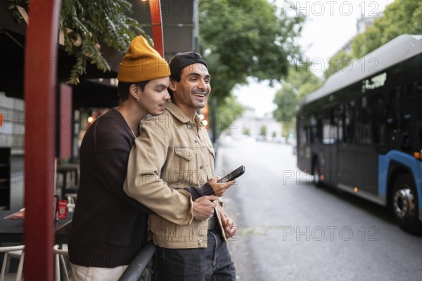 A joyful gay couple embraces by a bus stop. They are smiling, holding a phone, with trees, buildings, and a bus in the background, conveying a sense of warmth and connection