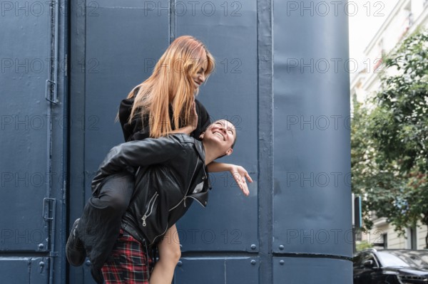 A joyful lesbian couple shares a playful piggyback ride in an urban setting, laughing and enjoying each other's company against a backdrop of blue and greenery