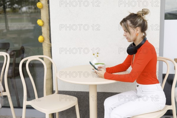A woman in a vibrant orange top sits at an outdoor cafe in Warsaw, using her smartphone with headphones around her neck The scene conveys a blend of leisure and technology