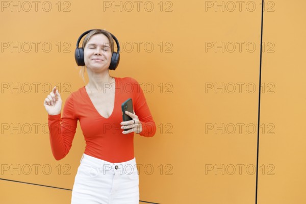 A woman stands against an orange wall in Warsaw, wearing headphones and a red top She holds a smartphone and seems to be enjoying music, smiling with eyes closed