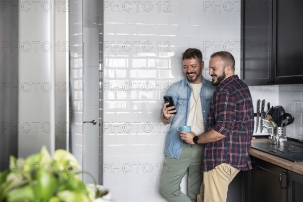 A happy gay couple stands in a bright kitchen, sharing a joyful moment while looking at a smartphone together. One holds a mug, creating a cozy and relaxed atmosphere