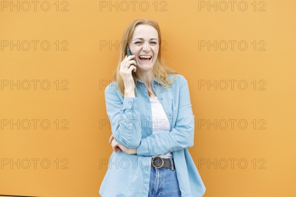 A cheerful woman in a blue shirt talks on her smartphone against an orange background in Warsaw, Poland Her casual attire and wide smile convey a sense of joy and connection
