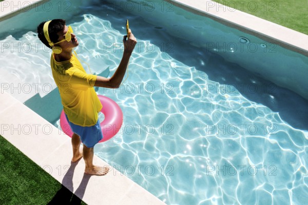 A young teenage boy in vibrant yellow attire takes a selfie by a sparkling blue pool. Sunlight reflects off the water's surface, highlighting the playful summer atmosphere