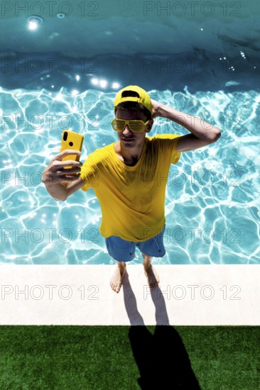 A teenage boy in a yellow shirt, sunglasses, and cap takes a selfie by a bright blue pool. His casual, colorful attire and the sparkling water capture a vibrant summer vibe