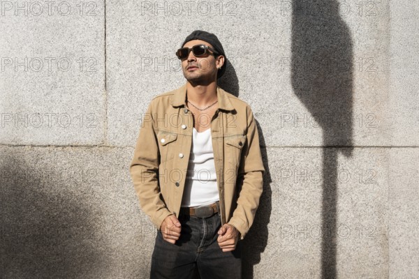 A man in a tan jacket and sunglasses leans against a sunlit concrete wall. The casual attire and shadow create a stylish, modern urban look