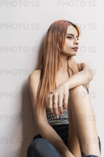 A woman with long hair sits against a white wall, looking contemplative. The close-up captures her relaxed posture and thoughtful expression