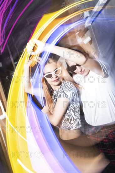A vibrant party scene features a dynamic lesbian couple dancing energetically, surrounded by swirling, colorful lights. The lively atmosphere is filled with motion and joy