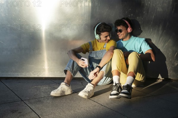 A gay couple sits joyfully against a metallic wall, sharing music through headphones. Their cheerful expressions and casual outfits capture the essence of togetherness and happiness