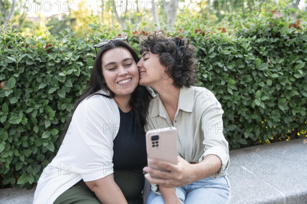 A joyful lesbian couple takes a selfie in a lush park. One partner kisses the other on the cheek, capturing a romantic moment filled with love and happiness