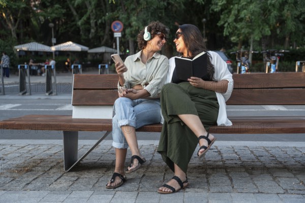 A lesbian couple enjoying a sunny park day. One listens to music with headphones and holds a phone, while the other reads a book. The background features a bustling city setting