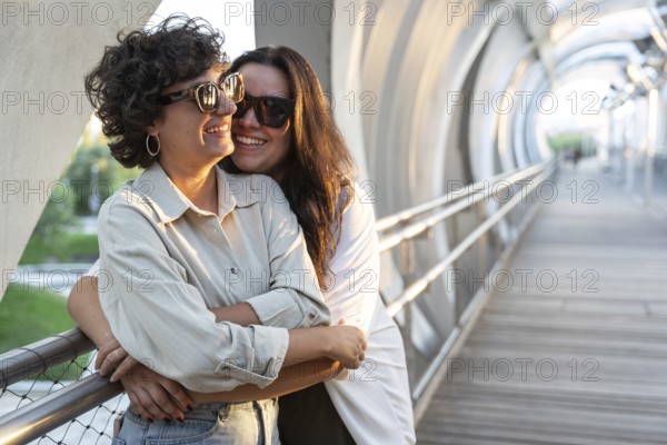 Lesbian couple warmly embracing while standing on a sunlit street bridge. Both wearing sunglasses, sharing a joyful moment. The scene captures love and happiness