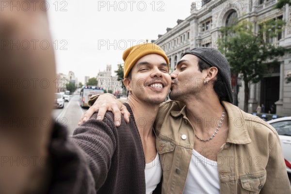 A joyful Colombian gay couple expresses affection on a bustling city street. Captured in a candid moment, one partner kisses the other on the cheek, radiating happiness