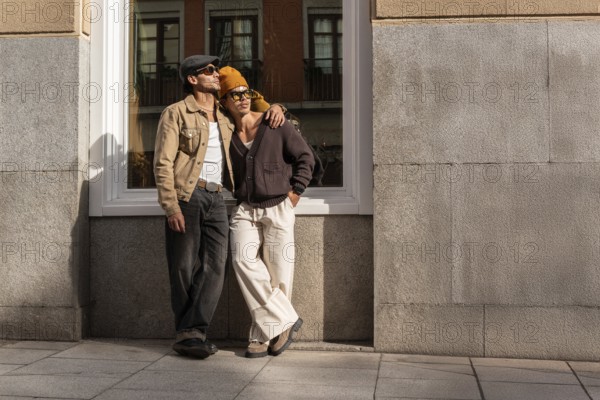 A Colombian gay couple in casual outfits enjoys a sunny urban setting. They lean against a building, embracing, reflecting affection and modern style