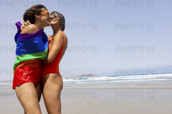 Two lesbians women embrace on a sunny beach, wrapped in a rainbow pride flag, symbolizing love and diversity. Wearing swimsuits, they enjoy a joyful, carefree moment by the ocean waves