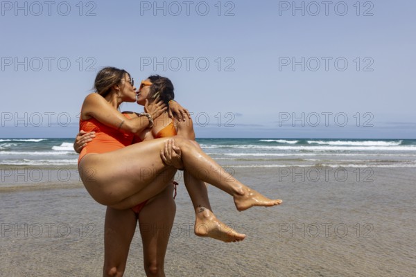 A lesbian couple in swimsuits shares a joyful embrace and kiss on a sunny beach, capturing a moment of love and happiness against the ocean backdrop. Perfect for themes of romance and summer