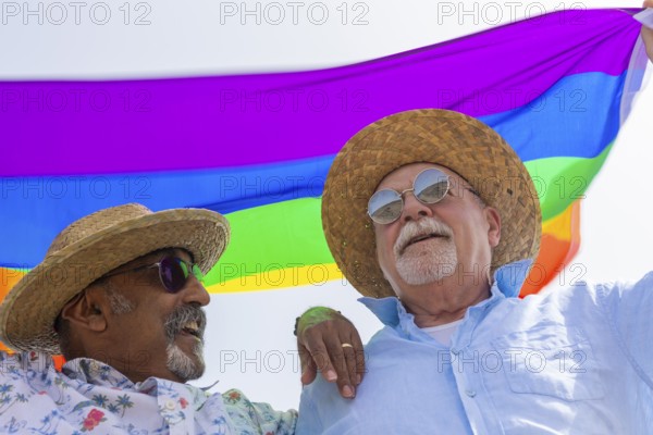 From below joyful gay senior couple, comprising two men, is captured outdoors, sharing a happy moment under a colorful rainbow flag. They are both wearing hats and sunglasses, with one man smiling at the other