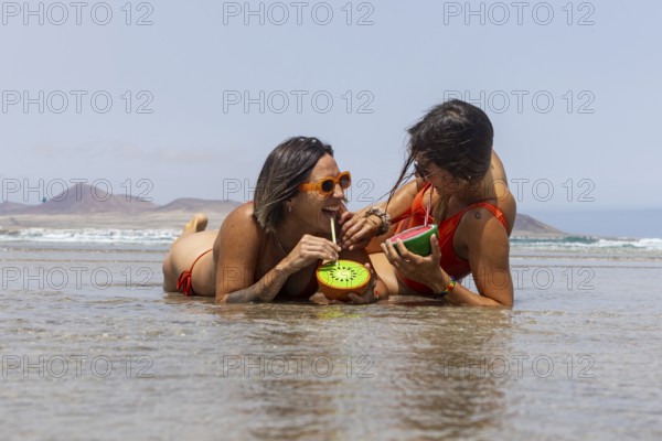 Two lesbians women in swimsuits enjoy refreshing drinks on the beach, laughing and relaxing by the water. The sunny day highlights their summer vibes