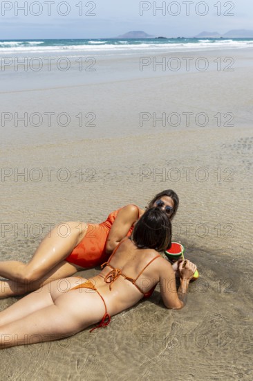 Couple of women in swimsuits lie on wet sand by the ocean, sharing a drinks under the sun. The scene captures a perfect summer day