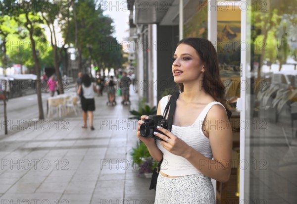 A young woman stands on a bustling city street, holding a camera Surrounded by passersby, she admires the urban landscape Trees and shops line the lively avenue