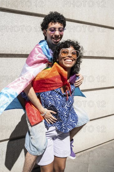 A vibrant, joyful multiethnic LGBTQ+ couple enjoys a sunny day outdoors, wrapped in pride flags. They are smiling and embracing, celebrating love and diversity in an urban setting