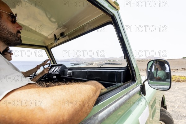 A blind man and a woman drive a motorhome along a coastal road with scenic mountains in the distance. The image highlights their adventurous spirit and connection to nature