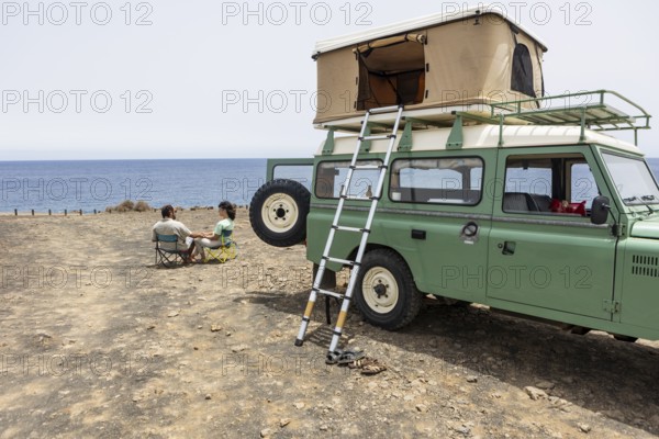 A blind man and a woman sit on camping chairs near their motorhome, overlooking the ocean. The rooftop tent and coastal backdrop highlight their adventurous spirit and relaxation