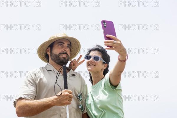 A blind man and a woman smile while taking a playful selfie by the sea. The woman makes a peace sign, capturing a joyful moment in their shared coastal adventure