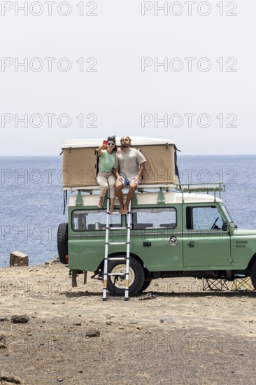 A blind man and a woman sit on the rooftop tent of a motorhome, enjoying a coastal view while taking a selfie. The scene reflects adventure, connection, and outdoor exploration