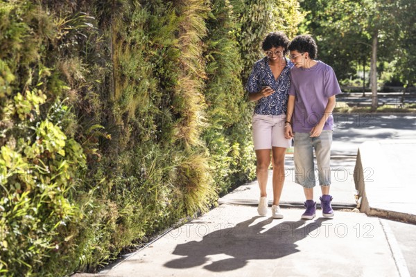 A loving multiethnic LGBTQ+ couple smiles as they enjoy a sunny day, walking side by side along a lush, green urban path