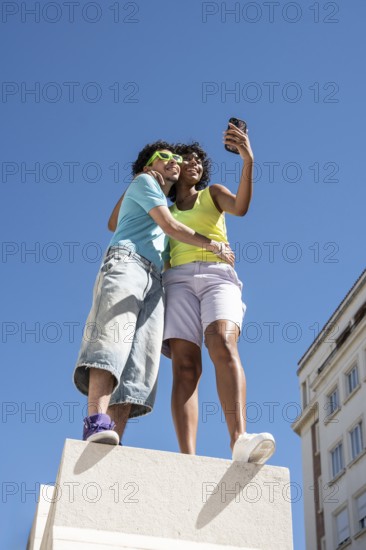 A joyful, multiethnic LGBTQ+ couple captures a selfie outdoors. They stand closely on a sunny day, showcasing unity and happiness against a clear blue sky