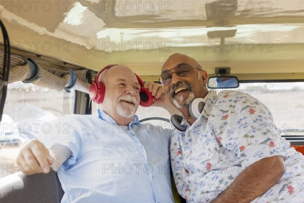 A senior gay couple smiles joyfully inside an off-road vehicle during a camping adventure. One man, bald with a white beard, wears red headphones, while the other, with a full grey beard, sports white headphones