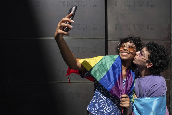 A joyful multiethnic LGBTQ+ couple takes a selfie wrapped in a rainbow flag, symbolizing pride and love. They are smiling, capturing a moment of happiness and unity